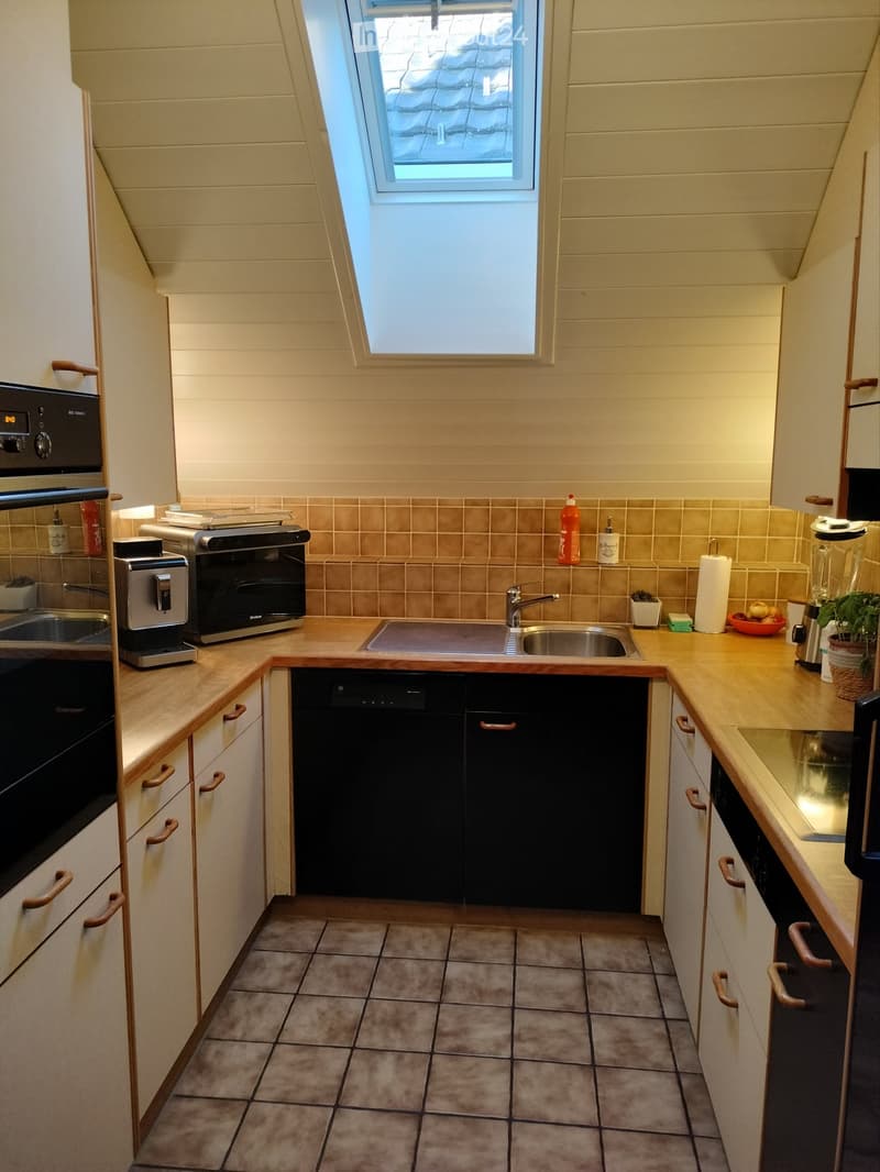 Modern kitchen with light wooden cabinets, stainless steel appliances, and a central island with a sink and faucet. The space features tiled flooring and a skylight window above the sink.