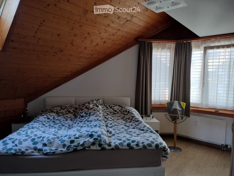 Cozy attic bedroom with wooden slanted ceilings, large windows with curtains, a double bed with patterned bedding, a nightstand, and a clothing rack.