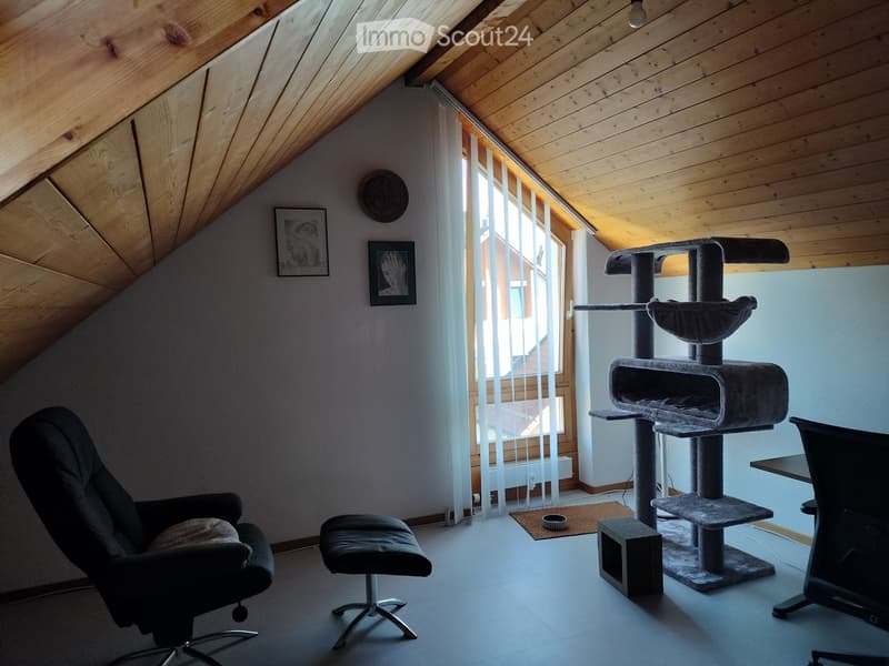 Cozy attic room with slanted wooden ceiling, large window with white blinds, cat tree, black leather chair, small stool, and a table.
