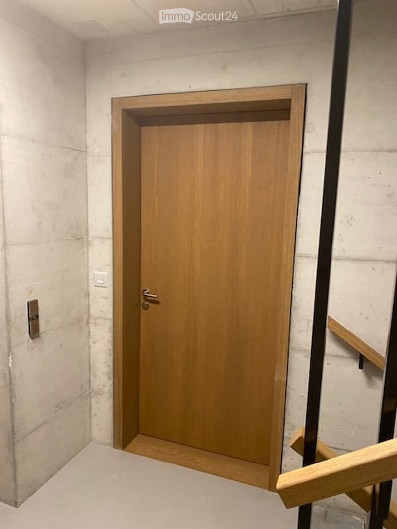 A narrow hallway with a wooden door and a staircase railing visible. The walls are concrete, and the floor is tiled.