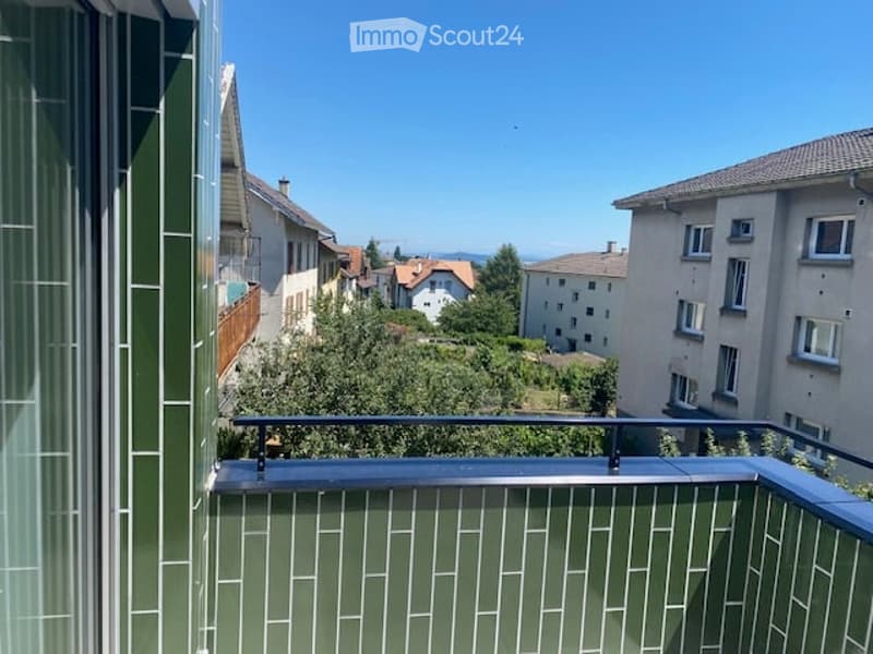 Balcony with metal railing and green tiled walls, overlooking a residential area with houses, trees, and distant hills under a clear blue sky.