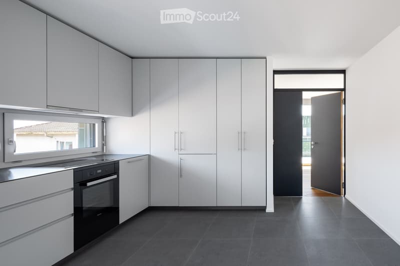 Modern kitchen with white cabinets, black oven, and dark tiled floor. Features a large window and an open doorway leading to another room.