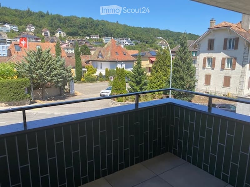 Balcony with a view of a town, featuring tiled flooring and a metal railing, overlooking a street with parked cars and surrounding buildings.