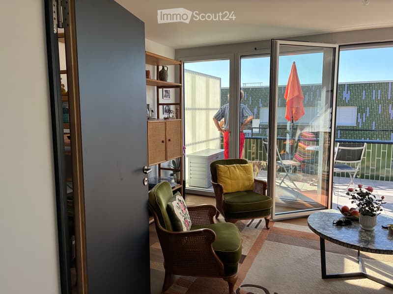 Bright living space with large glass doors opening to a balcony, featuring wicker chairs, a round table, shelves, and a cabinet.