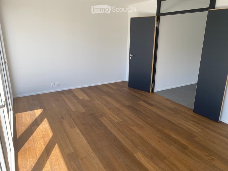 Empty room with light-colored walls and wooden flooring, featuring a closed door and a partially visible window frame.