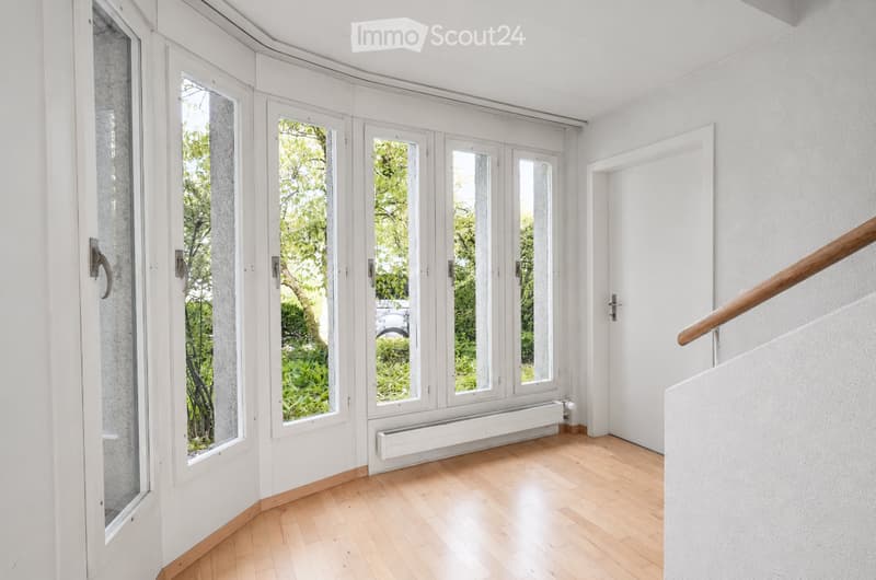 Bright hallway with wooden flooring, large windows, and a staircase with a wooden handrail.