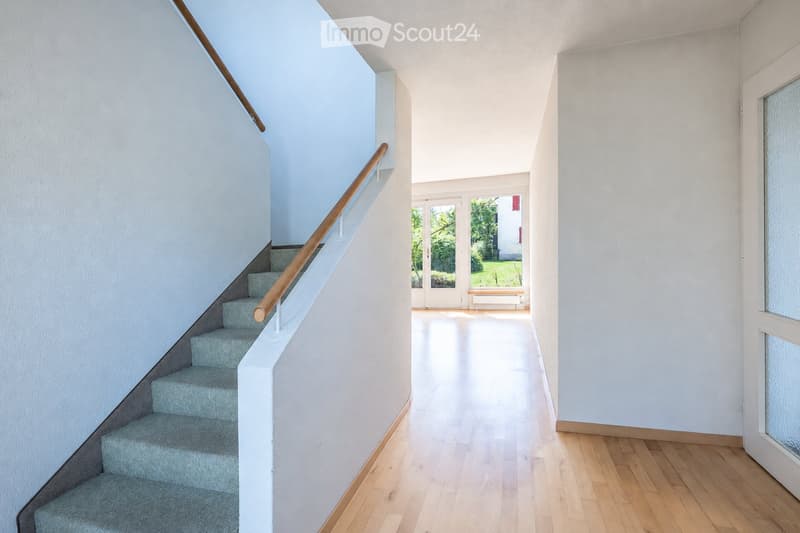 Bright hallway with wooden flooring, a staircase with a wooden handrail, and a view to an outdoor area through a glass door.