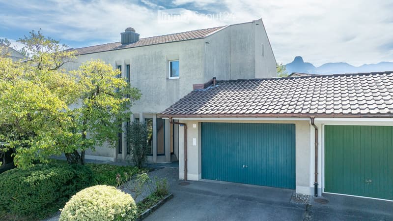 Two-story house with beige exterior, tiled roof, and green garage doors. The house features a well-maintained garden with shrubs and trees, and a driveway leading to the garage.