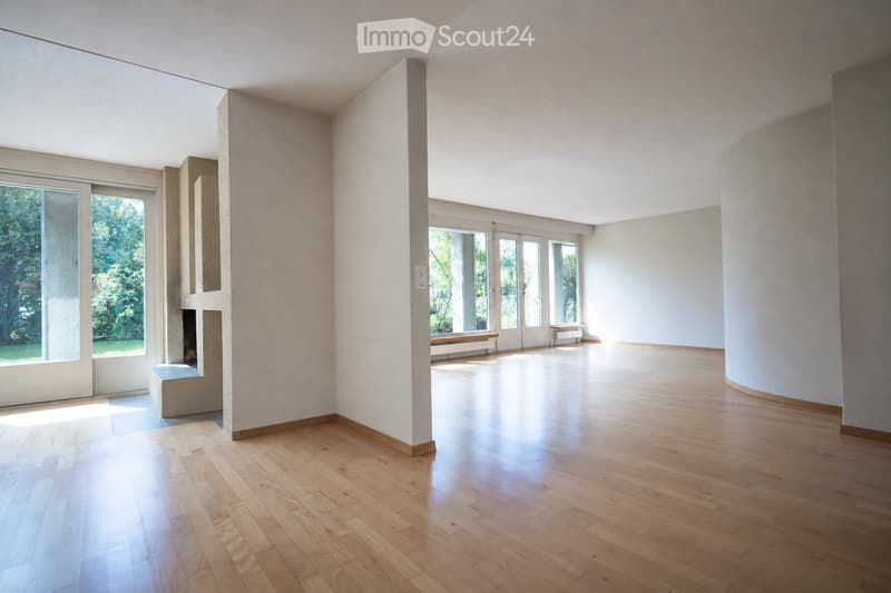 An empty, minimalist room with light wooden flooring, white walls, and large windows allowing natural light to flood the space. The room features a fireplace on one side and a bench in the background.