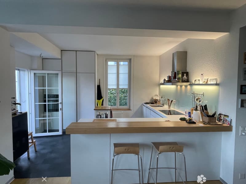 Modern kitchen with white cabinets, wooden bar counter, two stools, sink, oven, and various kitchen items on the counter and shelves.