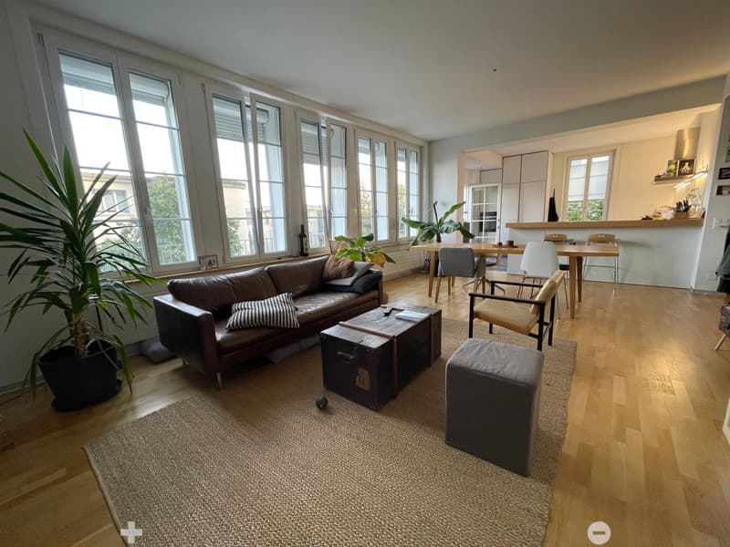 Modern living room with large windows, leather sofa, coffee table, ottoman, dining area with table and chairs, and plants.