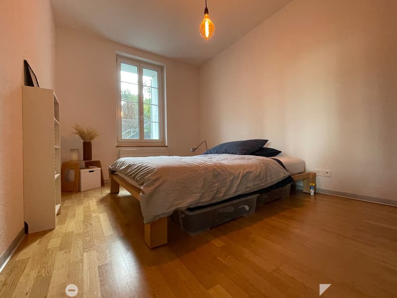 Cozy bedroom with wooden flooring, large window, bed with pillows and blankets, nightstand, and decorative vase.