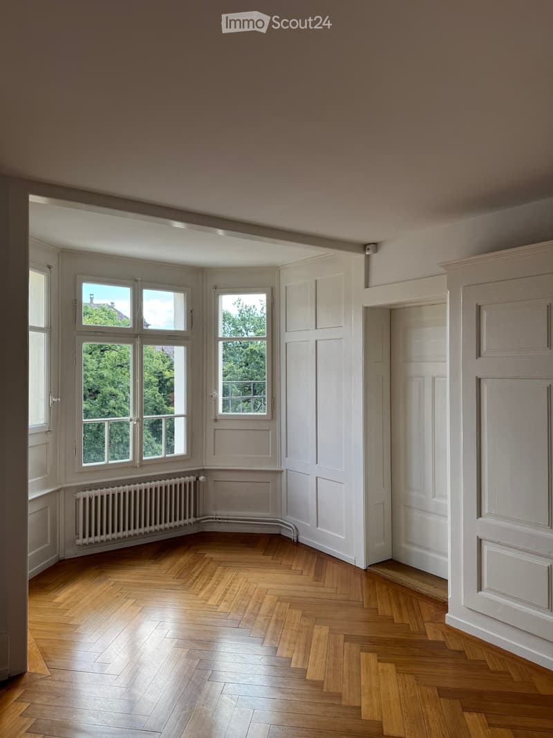 Empty room with wooden herringbone flooring, white walls, large windows with views of greenery, and a radiator under one window.