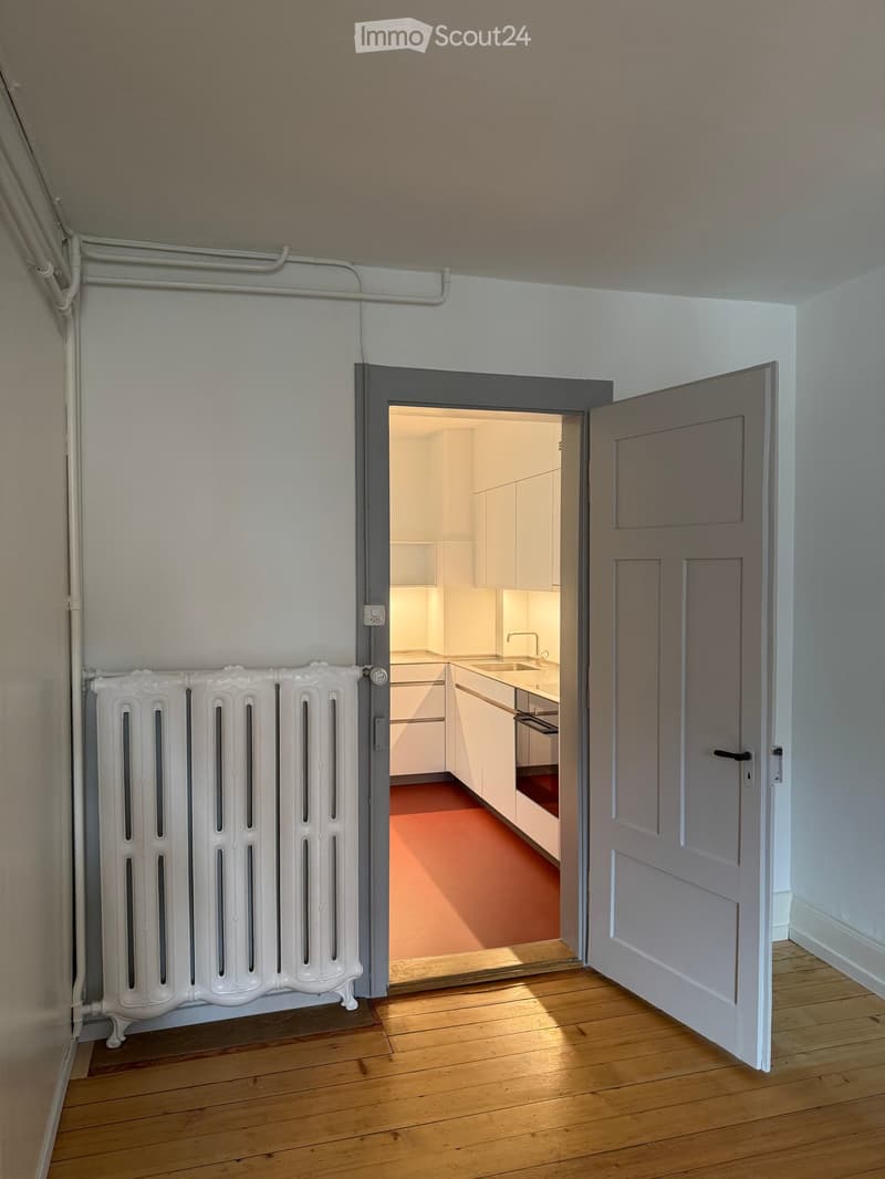 A narrow hallway with wooden flooring, white walls, and a radiator on the left side. An open door leads to a modern kitchen with white cabinets, a sink, and a countertop.