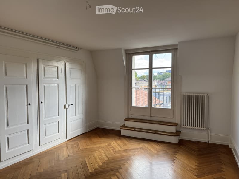Empty room with wooden herringbone flooring, white walls, three white doors, a window with a view of rooftops, a radiator, and a small step platform near the window.