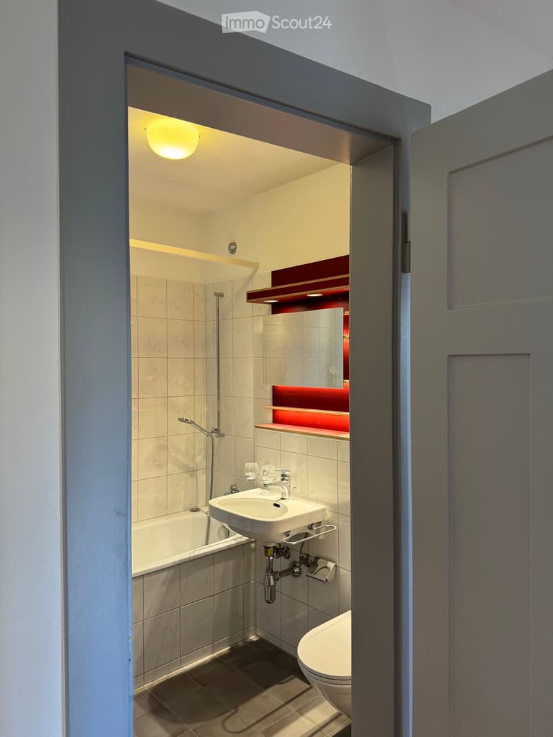 Modern bathroom with a white sink, toilet, bathtub, and a red shelf above the mirror. The room features beige tiles and warm lighting.