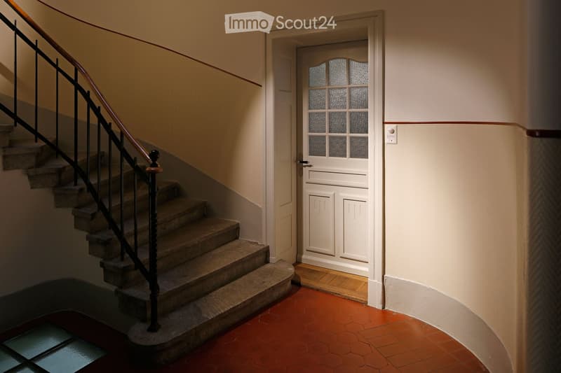 A narrow hallway with a staircase leading upwards, featuring a wooden handrail and a white door with frosted glass panels at the end.
