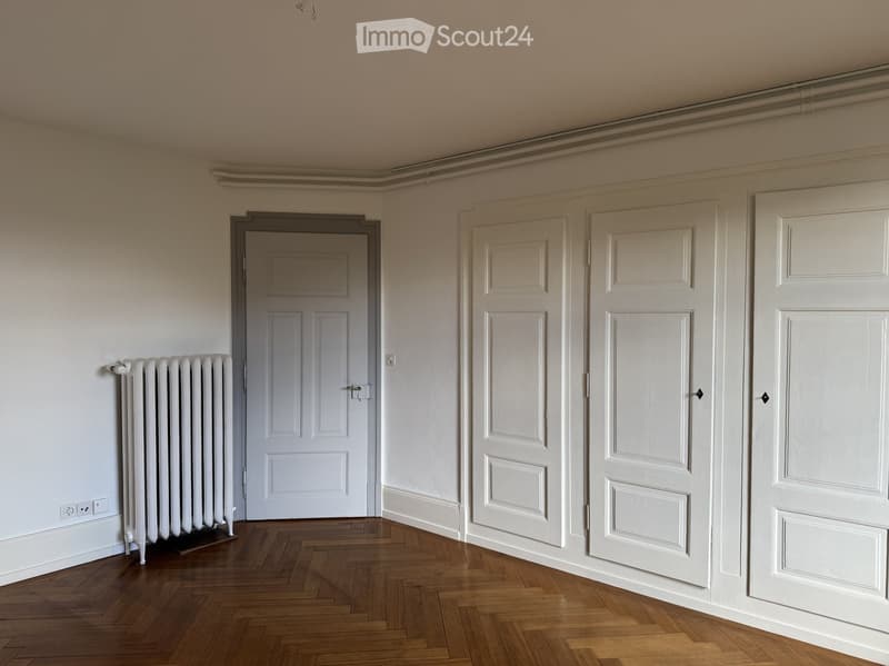 Empty room with wooden flooring, white walls, and several white doors leading to other spaces. A radiator is visible on the left wall.
