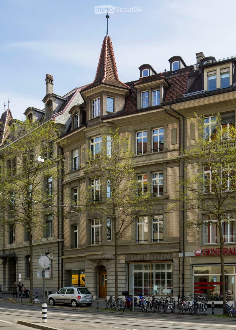 Multi-story building with ornate architecture, red-tiled roof, multiple windows, and balconies. The ground floor features storefronts with glass windows and doors. Bicycles are parked along the sidewalk, and a small car is visible on the street.