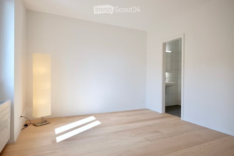 Bright, unfurnished room with light wooden flooring, white walls, and a tall floor lamp near the corner. A doorway leads to a small, modern bathroom with white tiles and a sink.