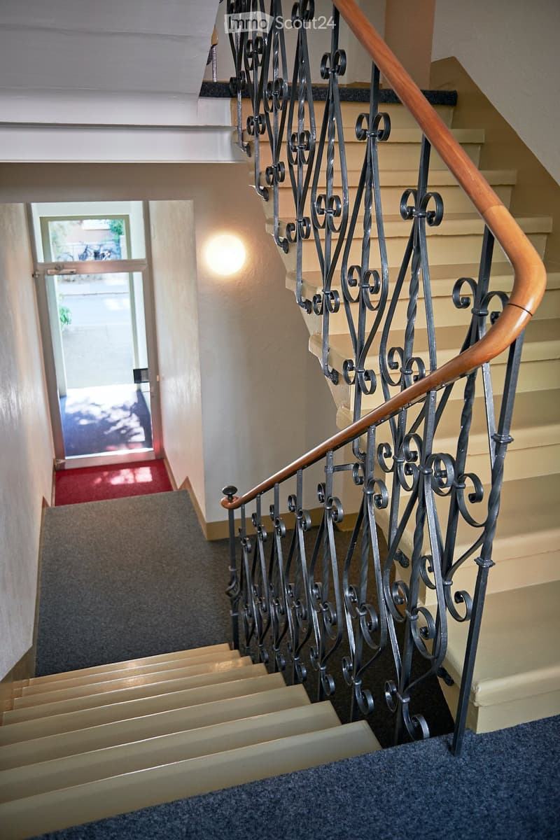 Staircase with ornate wrought iron railing and wooden handrail, carpeted steps, and a lit wall sconce.