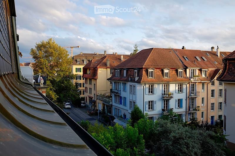 A view of a residential neighborhood with multi-story houses, red-tiled roofs, and balconies. The street below has parked cars and trees.
