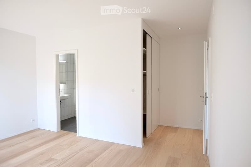 Empty room with light wooden flooring and white walls, featuring an open closet and a doorway to a small bathroom.