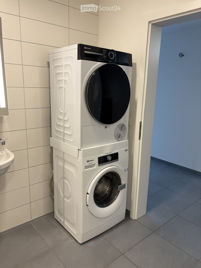 A white stacked washing machine and dryer. The washer on bottom, the dryer on top. Next to a white sink.
