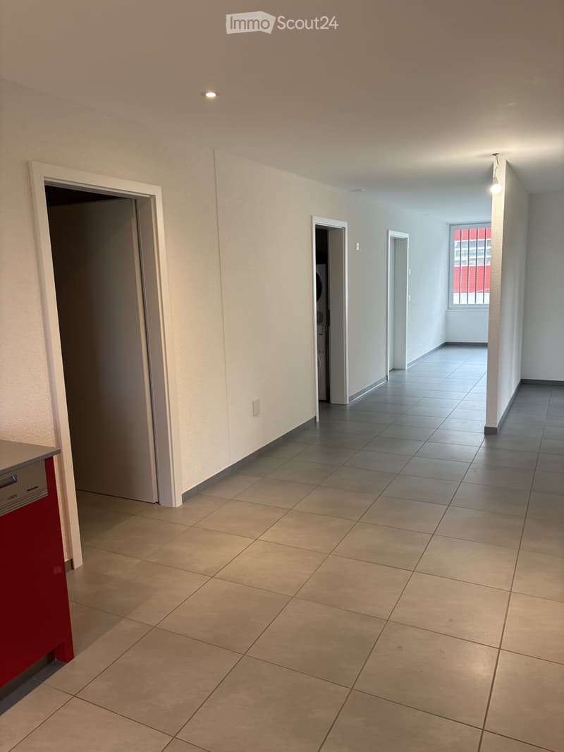 Empty room with tile flooring, white walls, and open doors. A red kitchen counter is on the left side. There is a closed white door on the right side, with an open door on the left. There is a lamp on the ceiling and a metal grill window.