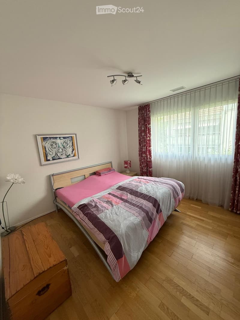 Bedroom with wooden flooring, a bed with pink and grey bedding, a nightstand with a lamp, a picture frame on the wall, and a window with curtains.