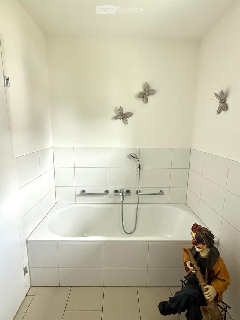 Modern bathroom with a white bathtub, chrome faucet, showerhead, and handrail. Decorative butterfly wall art is visible above the tub.