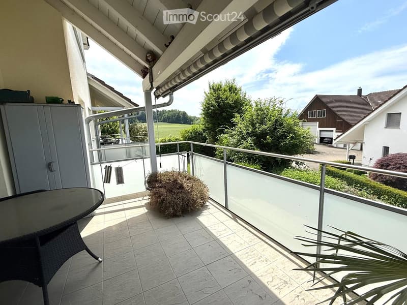 Balcony with tiled floor, glass railing, table, chair, and decorative plants, overlooking a residential area with houses and greenery.