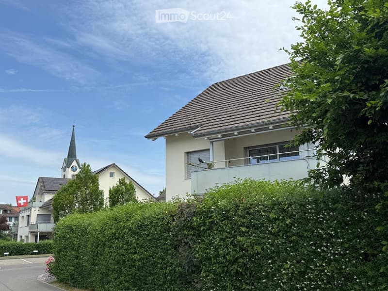 Two-story house with a tiled roof, white exterior, and a balcony on the upper floor. The house is surrounded by greenery and located near a church with a spire.