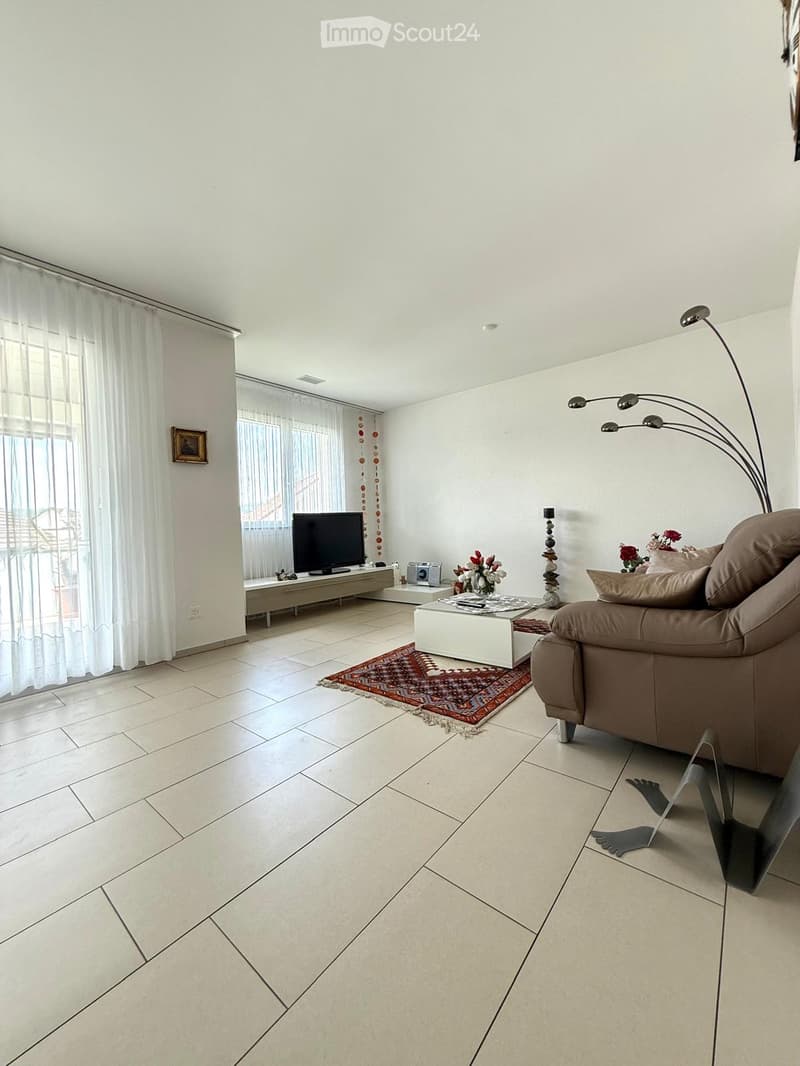 Bright living room with white walls, tiled floor, large windows with sheer curtains, a brown leather sofa, a white coffee table, a television on a stand, and decorative items including a flower vase and a picture frame.