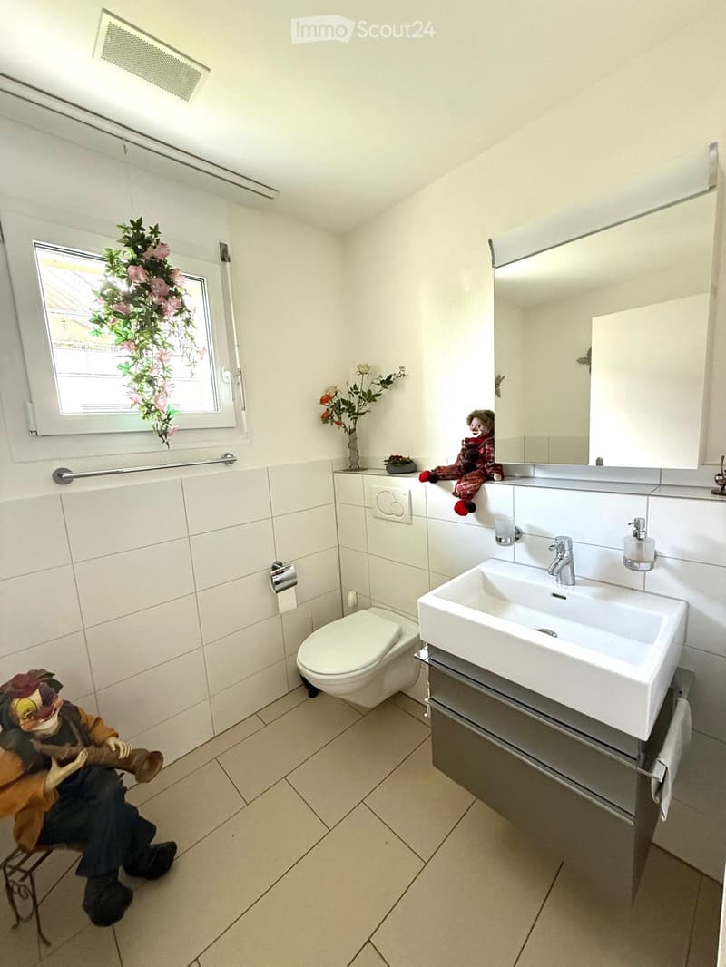 Modern bathroom with white tiles, a large sink with a modern faucet, a toilet, a mirror, and a small decorative stool with a clown figurine. A window with a floral decoration is visible.
