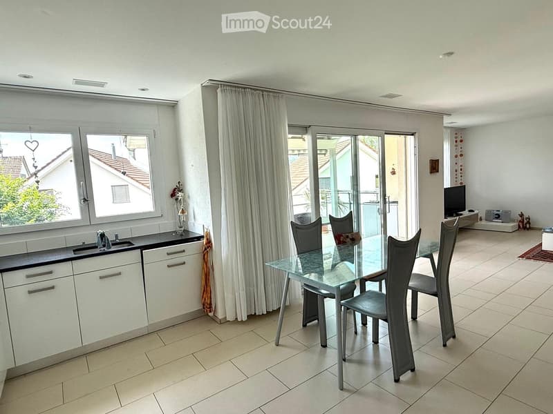 Bright, modern kitchen and dining area with white cabinets, a black countertop, a glass dining table with four chairs, and a TV in the background. Large windows and sliding glass doors lead to a balcony.