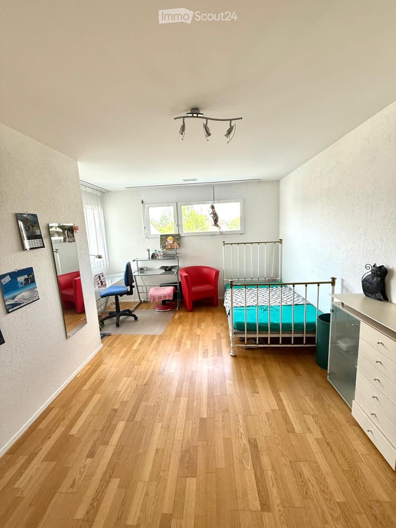 Bright bedroom with wooden flooring, a single bed with a metal frame and blue bedding, a red armchair, a desk with a chair, a mirror, and a cat statue on a drawer unit.