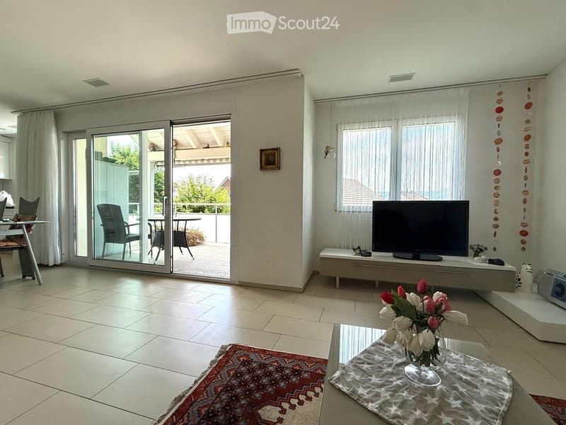 Bright living room with tiled floor, large sliding glass doors opening to a balcony, a television on a white console, a coffee table with a flower vase, and a patterned rug.