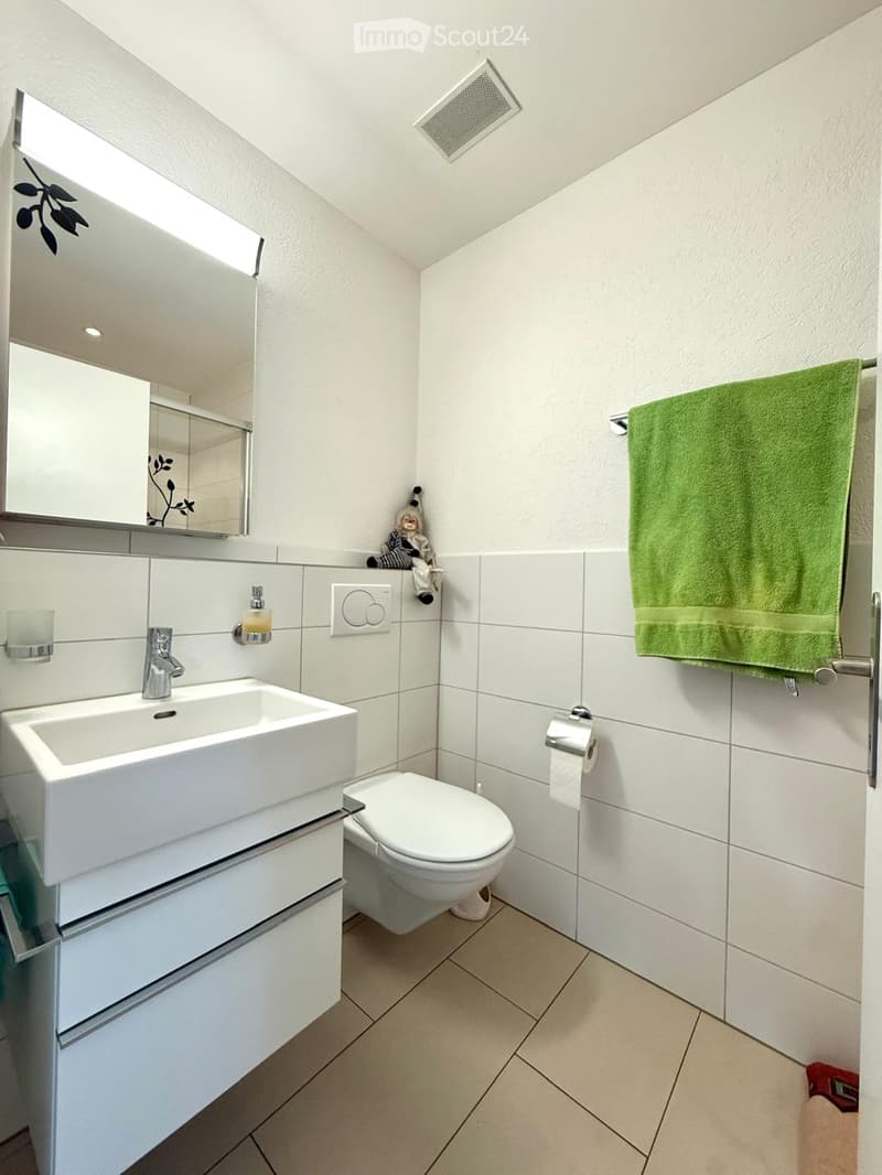Modern bathroom with white tiles, a large sink, toilet, mirror, and a green towel hanging on a rack.