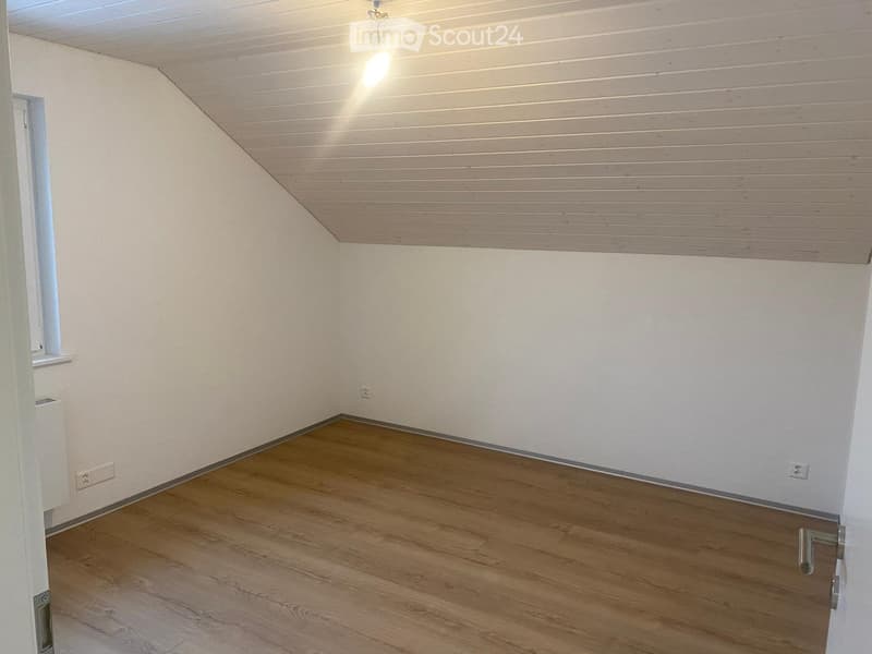 Empty room with wooden flooring, white walls, and a slanted wooden ceiling. Features a single light fixture and a small window with a shelf.