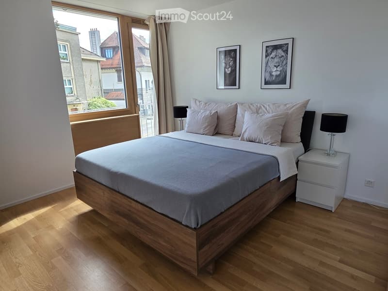 Cozy bedroom with a wooden bed, nightstand, lamp, and two picture frames on the wall. The room has wooden flooring and large windows with curtains.