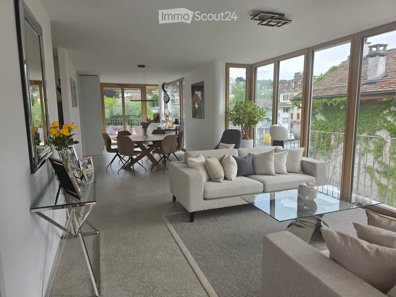 Modern living room with large windows, grey sofa, coffee table, dining area, and decorative items.