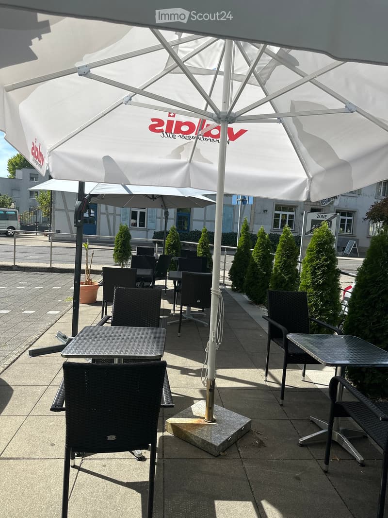 Outdoor seating area with black wicker chairs and tables under large white umbrellas, surrounded by neatly trimmed trees and a paved sidewalk.