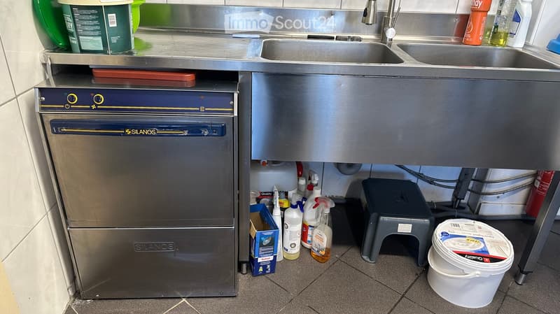 Commercial kitchen with stainless steel sink, faucet, oven, and various cleaning supplies on the floor and counter.