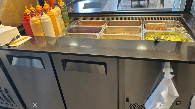 Stainless steel food preparation counter with multiple condiment bottles and serving trays, along with napkins and a pen on the counter.