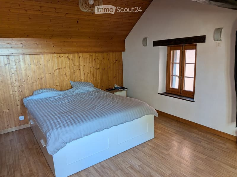 Cozy bedroom with wooden walls, slanted wooden ceiling, large window with wooden frame, and a neatly made bed with a checkered duvet.