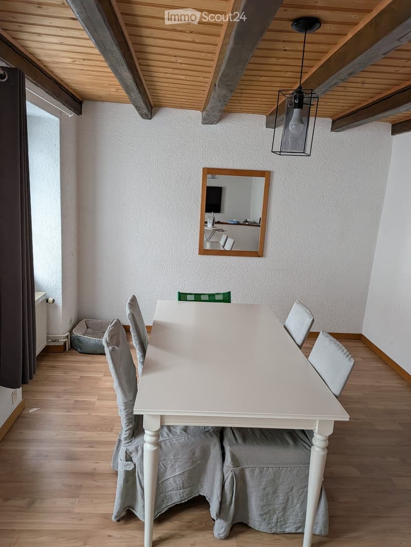 Dining area with a white table, four grey upholstered chairs, a mirror on the wall, and a hanging light fixture.