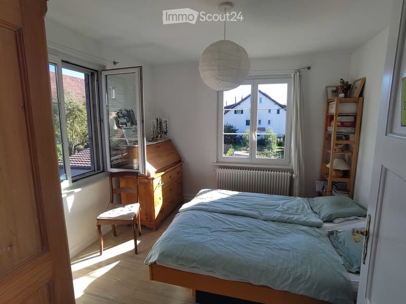 Cozy bedroom with a double bed, wooden chair, desk, shelf, and a hanging paper lantern light fixture. Large windows provide natural light and a view of the neighborhood.