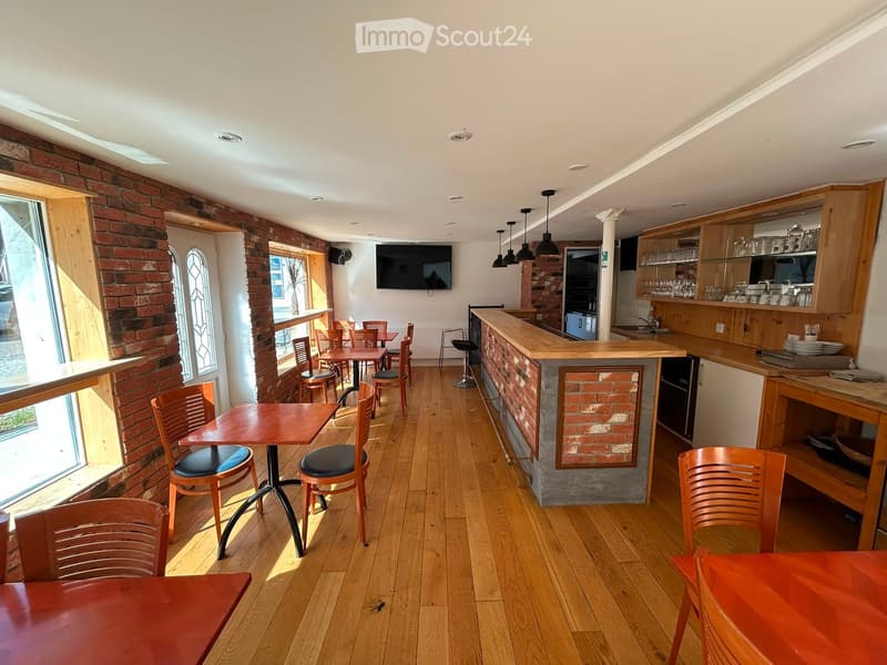 A cozy restaurant interior with wooden floors, brick walls, tables, chairs, a bar counter, and a television mounted on the wall.