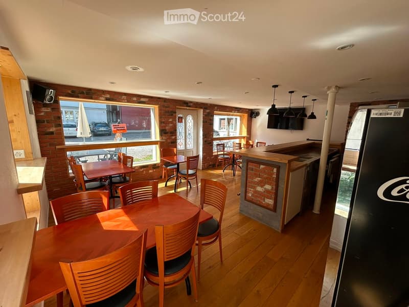 Restaurant interior with wooden tables and chairs, bar counter, refrigerator, and a television screen.